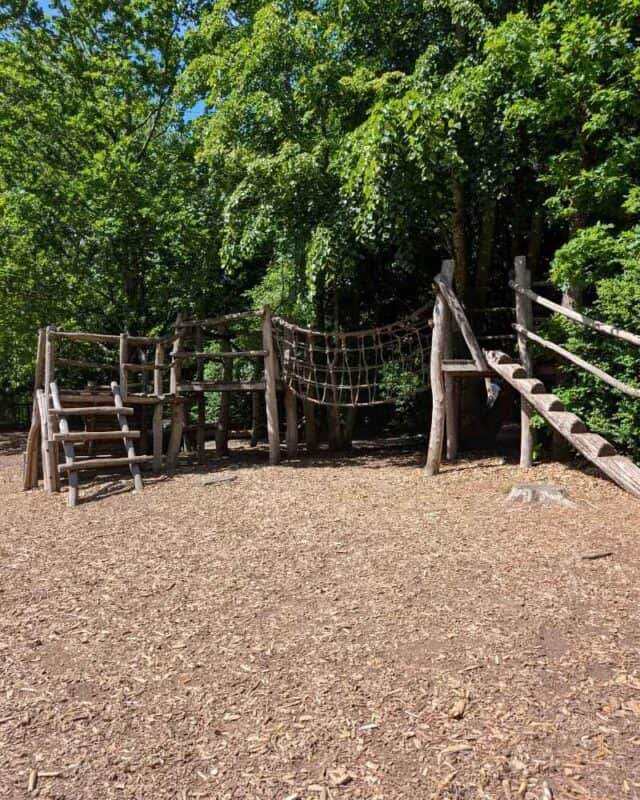 Natural woodland playground with rustic timber climbing frames, rope bridges, and balance elements set among mature trees. Woodchip surfacing covers the ground beneath the play equipment, creating an adventure play area that blends into the forest setting.