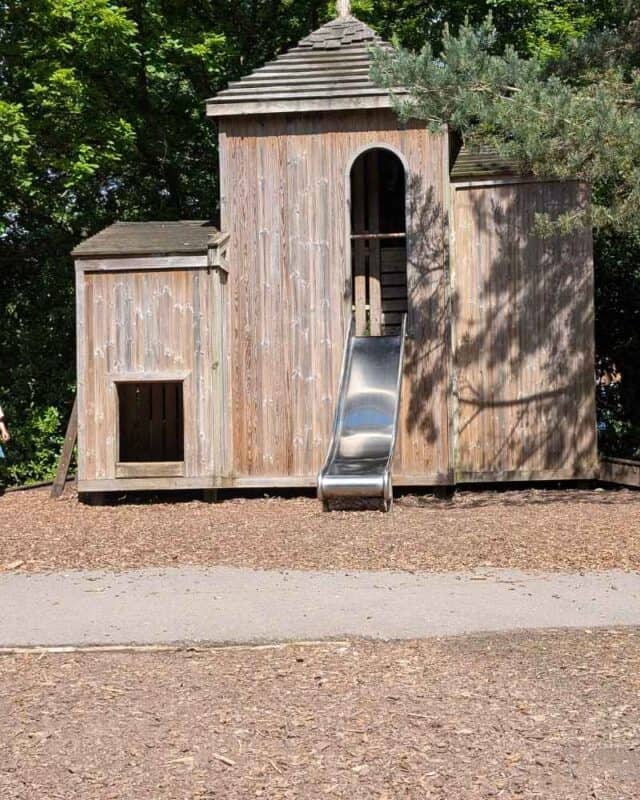 Wooden play tower with a metal slide and small crawl-through entrance in a woodland playground. The structure is surrounded by trees and woodchip surfacing, offering a simple climbing and sliding area for children.