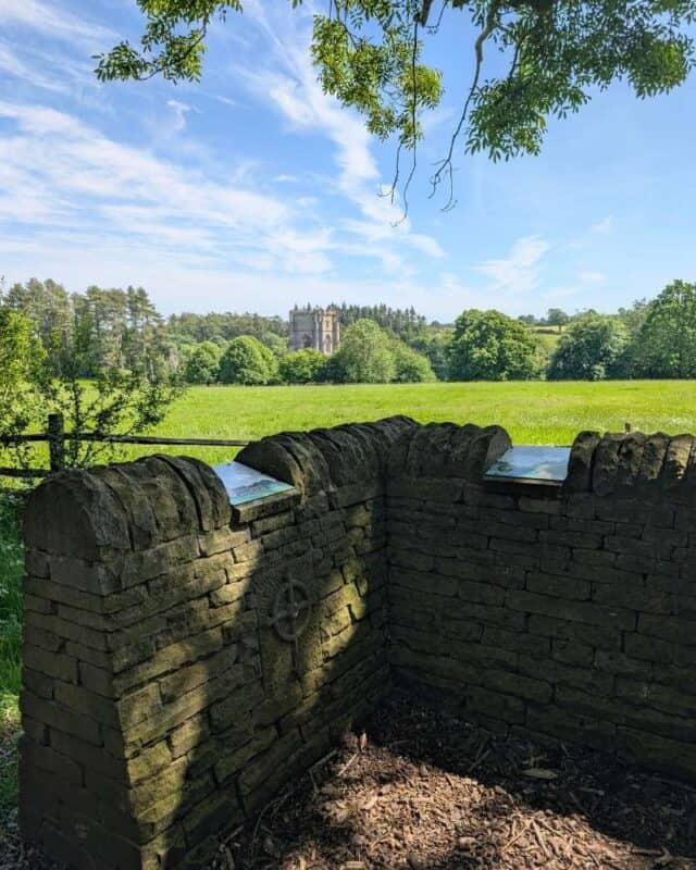 Stone viewing wall with interpretive panels overlooks a wide green meadow and a distant historic tower rising above the treeline. Overhanging branches frame the scene, while sunlight casts shadows across the weathered stonework in the foreground.