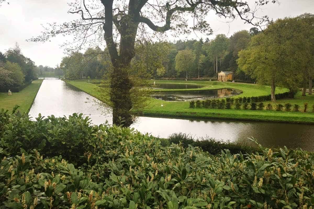 Formal landscaped garden with long rectangular canals and a circular pond surrounded by neatly trimmed grass and trees. A small classical pavilion stands beside the water, viewed through foreground shrubs and beneath the branches of a large tree.