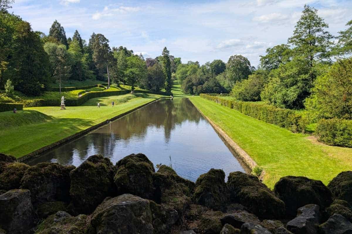 Long rectangular reflecting pool bordered by neatly trimmed lawns and hedges in a landscaped garden. Trees line both sides of the waterway, with a few visitors walking along the paths and statues visible in the distance.