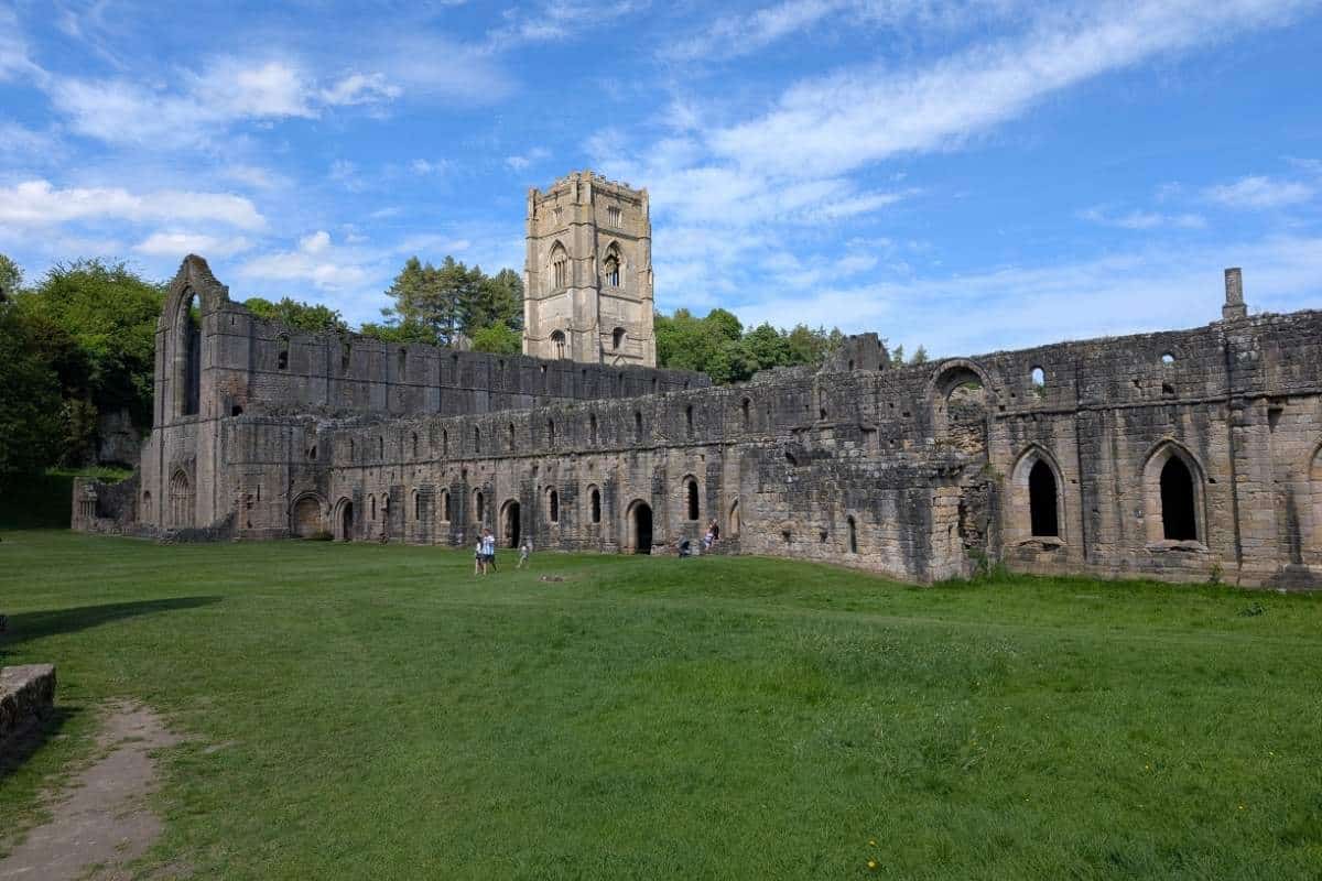 Wide view of Fountains Abbey ruins with long stone walls, arched windows, and the central tower rising above the monastery complex. Visitors walk across the expansive grassy grounds in front of the historic ruins beneath a blue sky with scattered clouds.