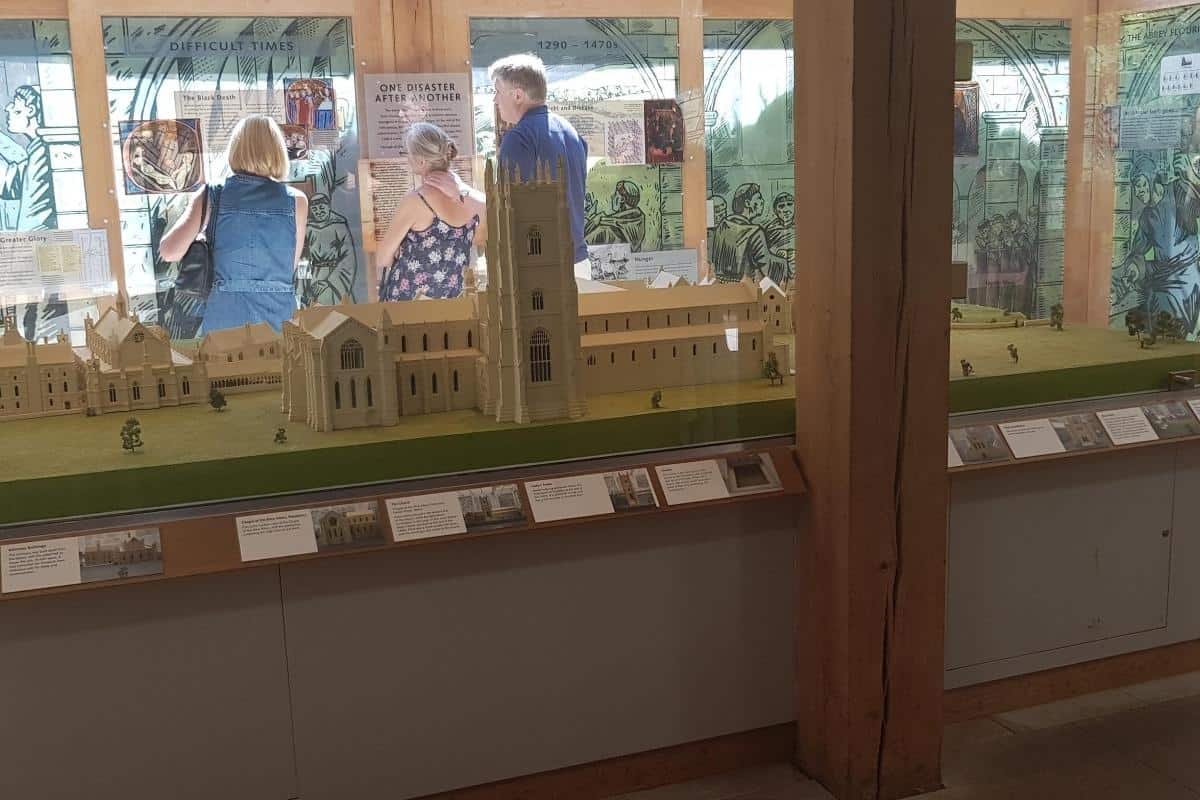 cale model of Fountains Abbey displayed inside a museum exhibit, showing the abbey church, central tower, and surrounding buildings as they would have appeared when complete. Visitors stand behind the display reading interpretive panels, including signs that read "Difficult Times" and "One Disaster After Another".