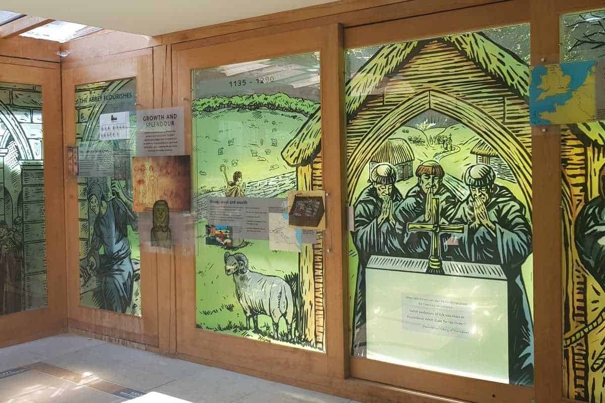 Museum exhibit panels illustrating the history of Fountains Abbey, featuring large drawings of monks praying, sheep grazing, and medieval rural life. Display text includes "Growth and Splendour" and dates "1135 - 1290", highlighting the abbey's period of prosperity and agricultural wealth.