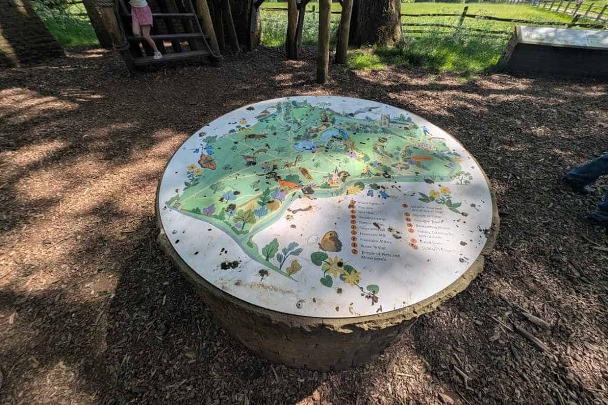 Circular illustrated map mounted on top of a tree stump in a woodland play area, showing landmarks, walking routes, wildlife, and attractions around the Fountains Abbey estate. Woodchip paths, a wooden climbing structure, fencing, and shaded trees surround the map, helping visitors explore the grounds.