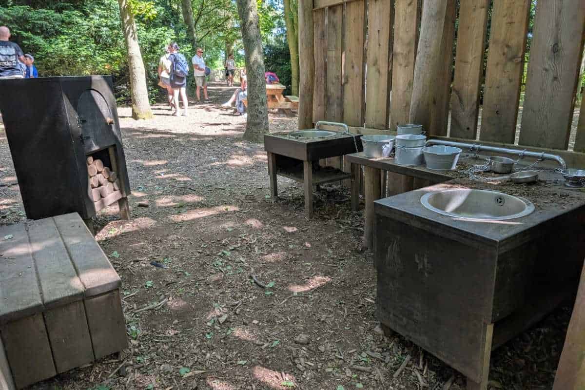 Outdoor woodland play area with child-sized metal sinks, buckets, and cooking stations set up beside a wooden shelter. Visitors walk along the shaded path in the background, while the mud kitchen encourages hands-on nature play among the trees.