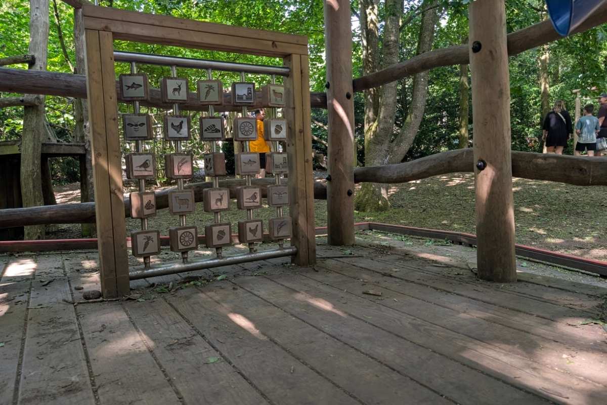 Wooden activity panel in a woodland adventure playground featuring rotating blocks illustrated with animals, plants, and nature symbols. The panel is mounted within a timber climbing structure, while children and adults explore the shaded forest play area in the background.