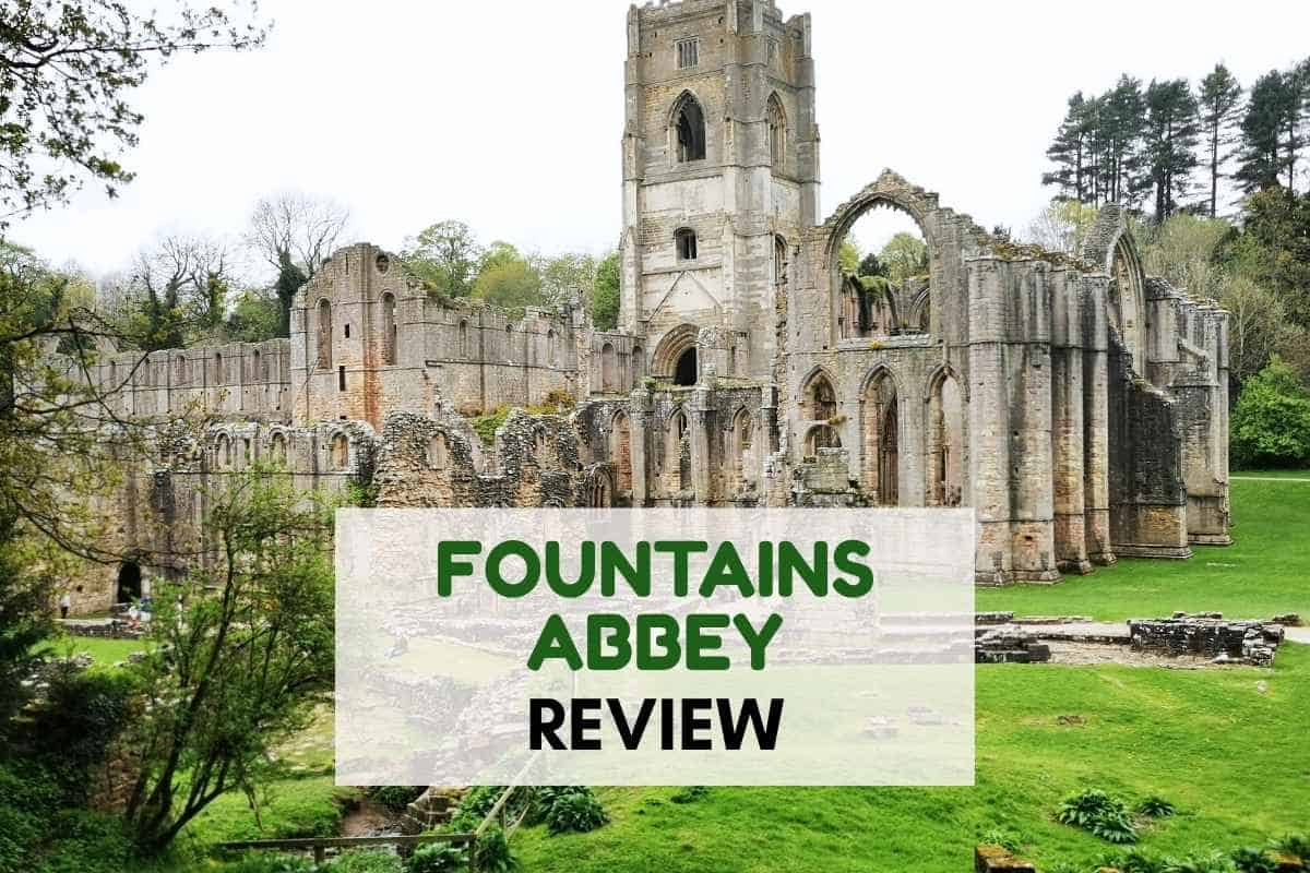 Ruins of Fountains Abbey viewed across a green lawn with stone arches and a tall central tower surrounded by trees. Overlay text reads "FOUNTAINS ABBEY REVIEW".