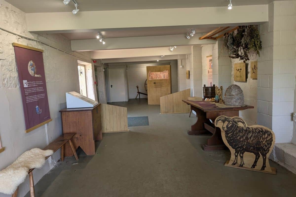 Museum-style exhibition space inside a historic building with information panels, wooden furniture, and interpretive displays. A table holds historical artifacts including a woven beehive-shaped skep, while a sheep illustration, dried herbs hanging from the ceiling, and benches line the room.