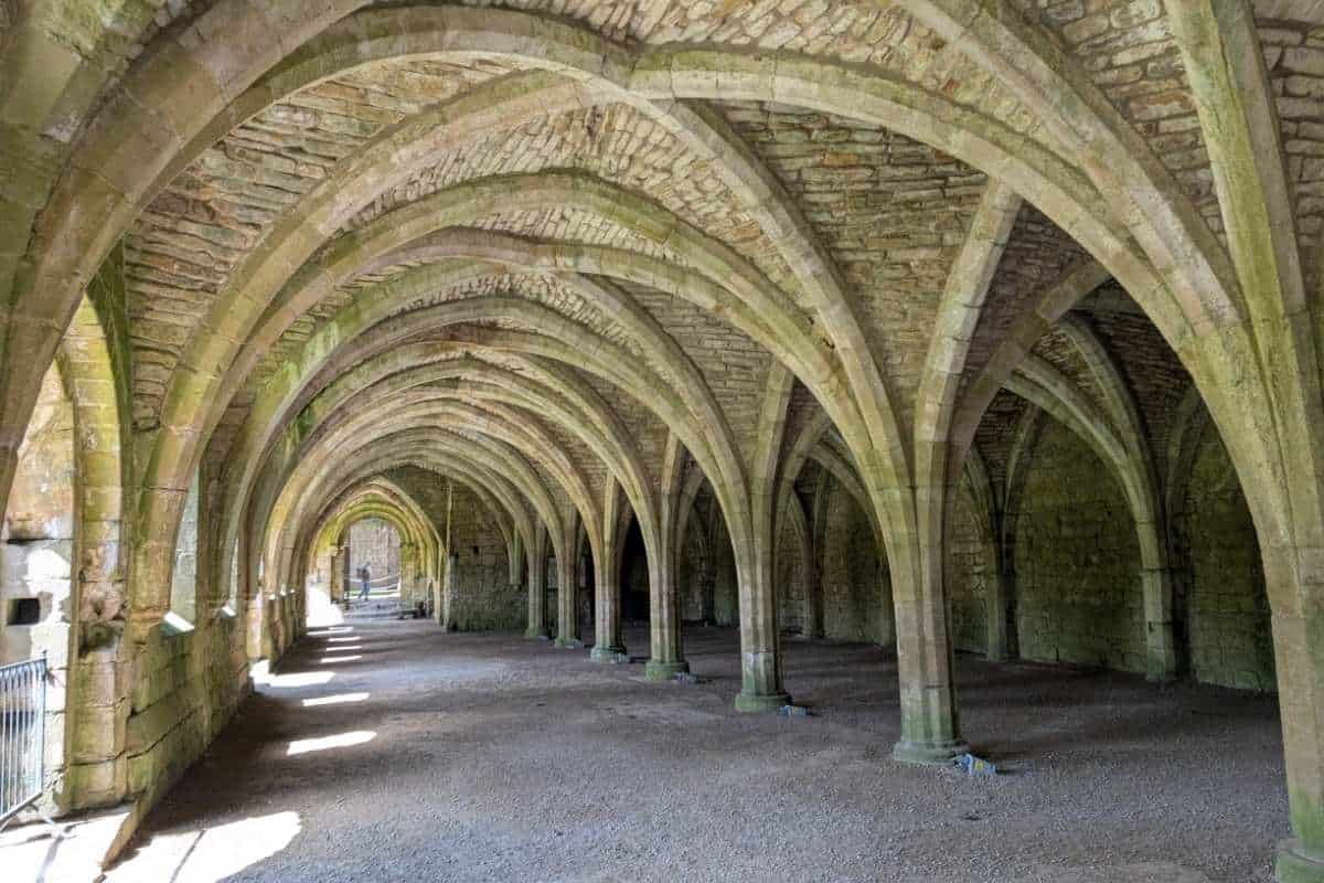 Long medieval stone hall with rows of pointed Gothic arches supporting a vaulted ceiling. Sunlight streams through openings along one side, creating bright patches on the floor and highlighting the repeating architectural details of the historic structure.