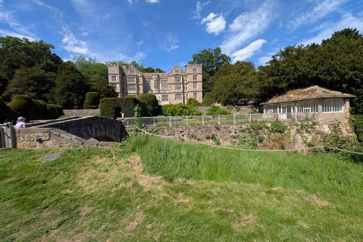 Historic stone manor house overlooking formal gardens with clipped hedges and terraces. A small stone bridge crosses a sunken garden area in the foreground, while a pavilion-like building stands to the right beneath a bright blue sky.