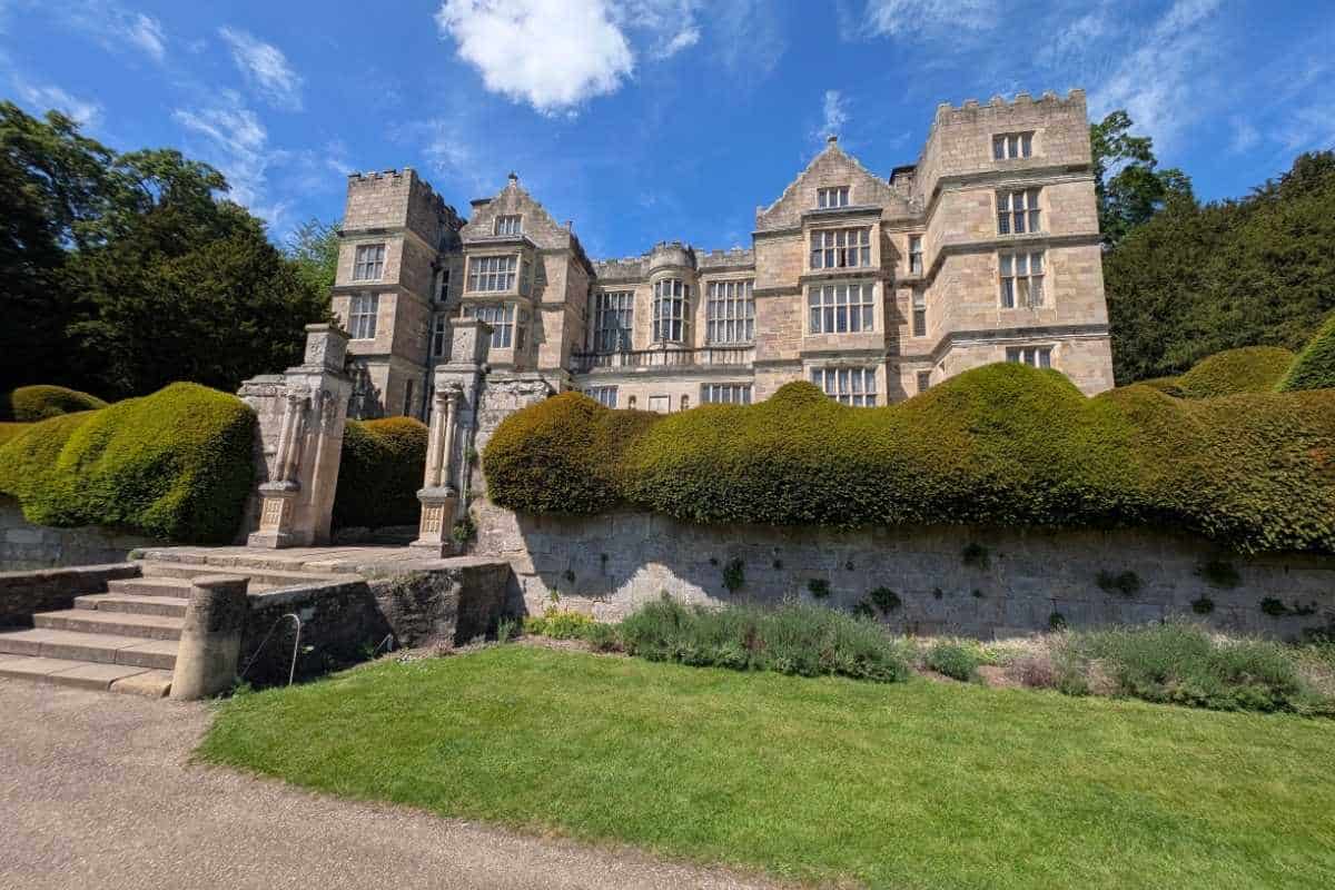 Large historic stone manor house with tall windows, decorative gables, and tower-like sections rising above clipped hedges. Stone steps and an ornate entrance structure lead up from a formal garden lawn under a bright blue sky.