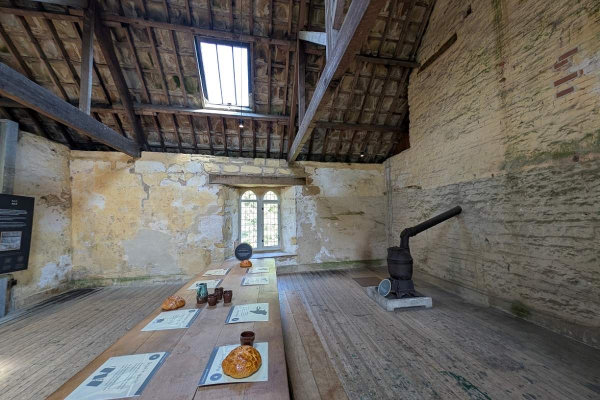 Historic room inside a restored abbey building with exposed timber roof beams, weathered plaster walls, and a tall arched window. A long interpretive table displays historical information and replica objects, while a cast-iron stove stands against the wall, illustrating daily life at the site.