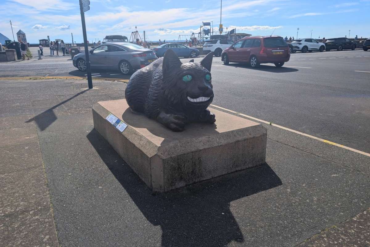 Black cat sculpture with bright green eyes and a wide grin displayed on Llandudno west shore promenade beside the seafront road. Cars, pedestrians, and seaside attractions can be seen in the background, making the statue a distinctive landmark along the town's waterfront.