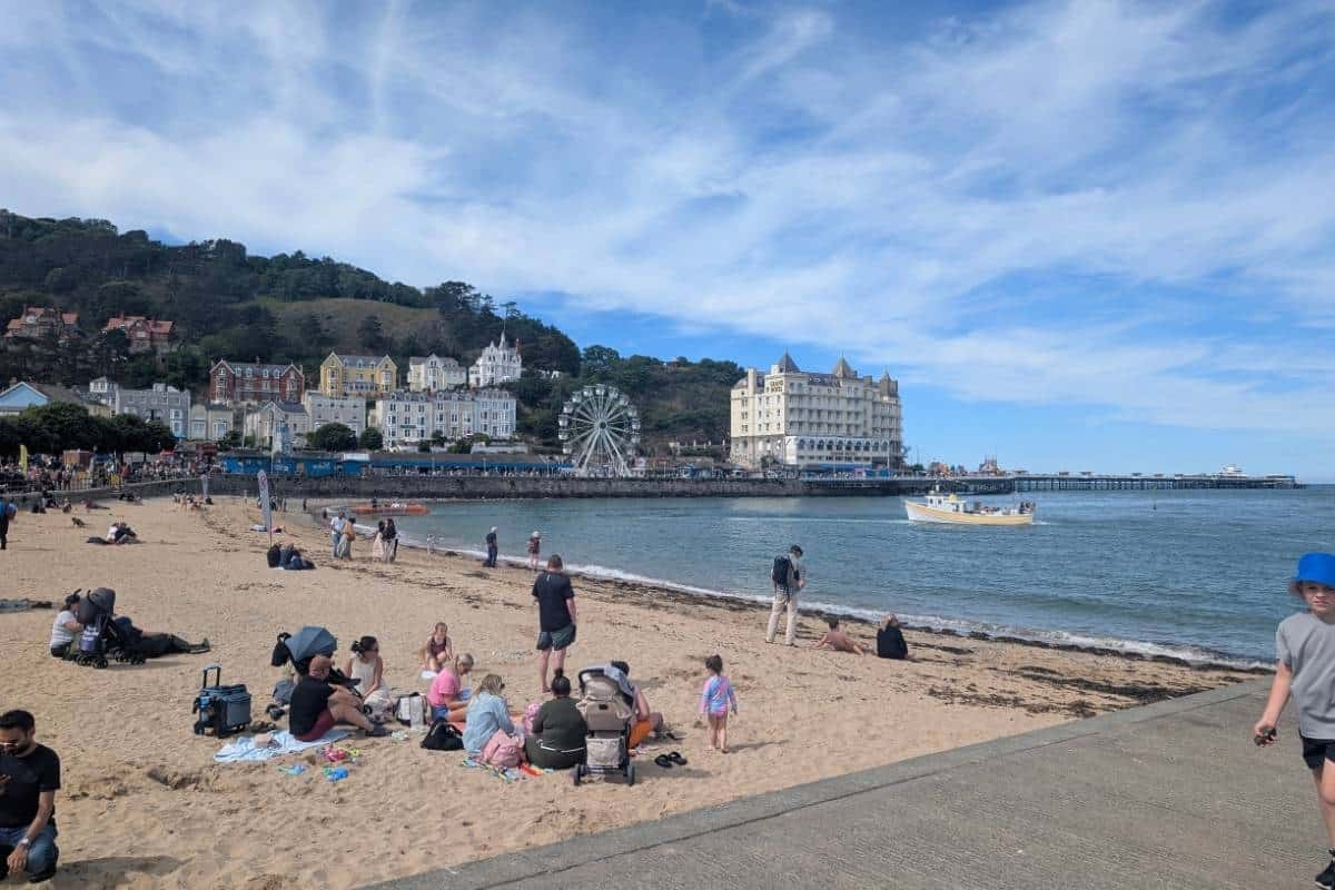 Sandy beach in Llandudno with families and visitors relaxing by the shoreline on a sunny day. The seafront features a large white hotel, a Ferris wheel, and the town's Victorian buildings backed by green hills, while a small boat floats on the calm sea near the pier.