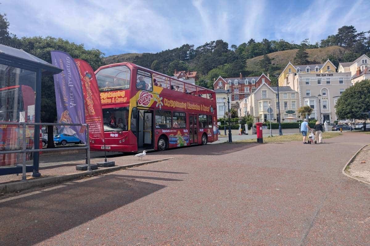 Open top sightseeing bus parked along the Llandudno promenade with passengers boarding for a coastal tour. Colourful Victorian buildings and green hills rise behind the seafront, highlighting one of the popular ways visitors explore the town and its surrounding scenery.