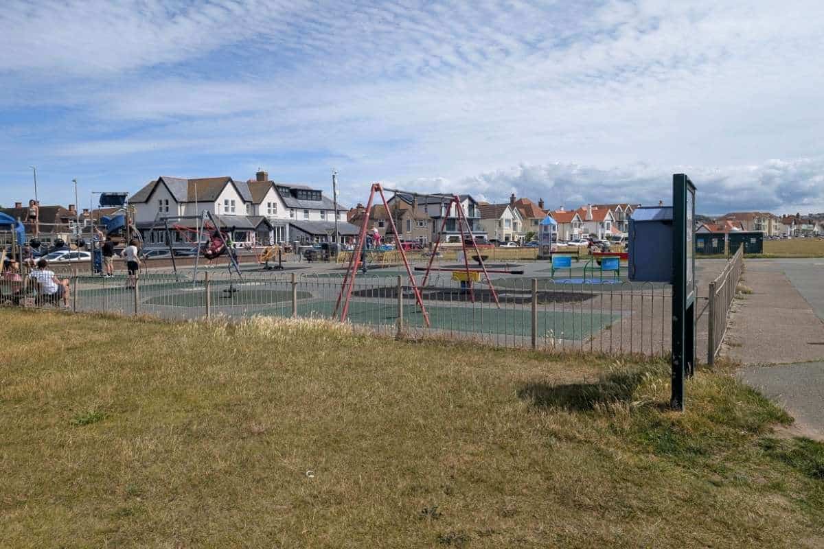 Alt text: Fenced playground in Llandudno with swings, climbing equipment, and children playing on a bright day. Residential houses and marina buildings line the background, highlighting a family friendly recreation area near the waterfront.