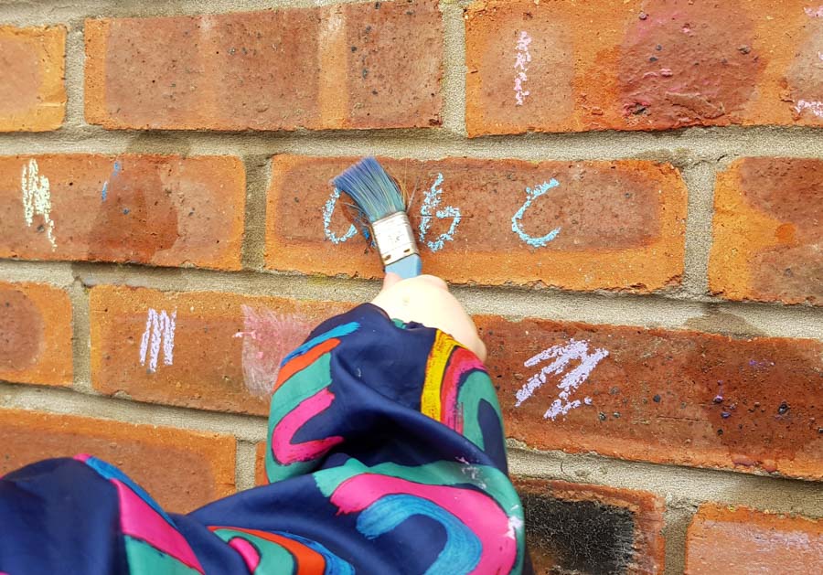 Toddler hand with a paint brush over chalk letters on a brick wall
