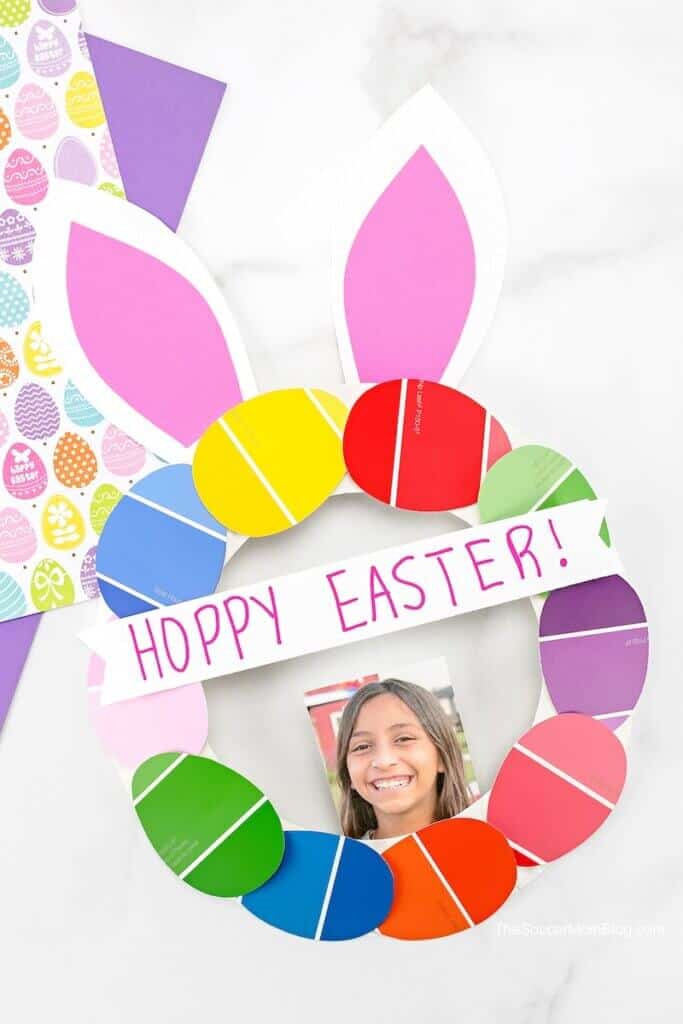 Paper wreath made of lots of egg shaped pieces of coloured card. It has bunny ears, a photo of a child and a banner that says Hoppy Easter