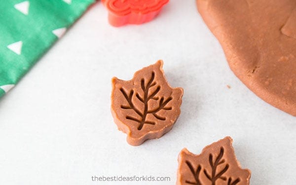 Brown playdough with leaf-shaped cutouts stamped using a red plastic leaf stamp on a white surface.