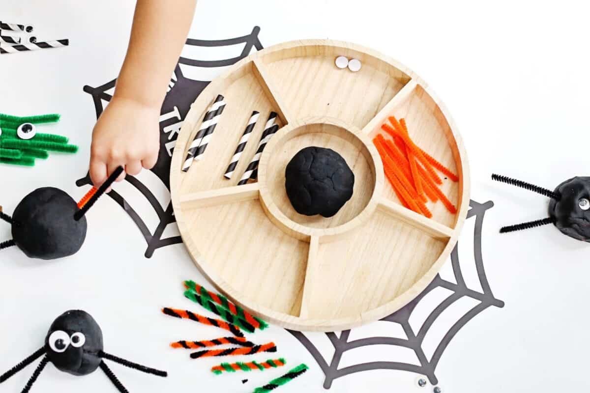 Round wooden tray with black playdough and different coloured straws and pipe cleaners and a little hand playing