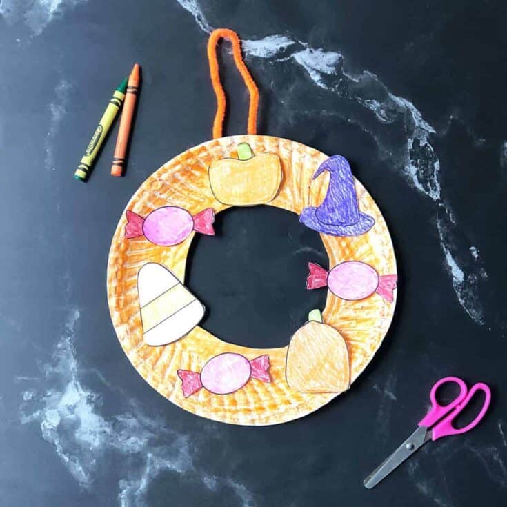 Halloween paper plate wreath craft decorated with hand-colored cutouts of candy, pumpkins, candy corn, and a purple witch hat, shown with crayons and scissors on a black background.