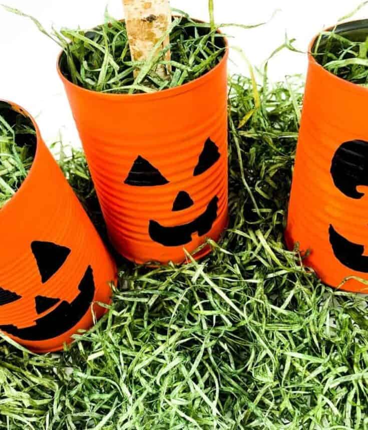 DIY Halloween craft with three orange tin cans painted as jack-o’-lanterns, filled with green shredded paper and sticks, placed on artificial grass