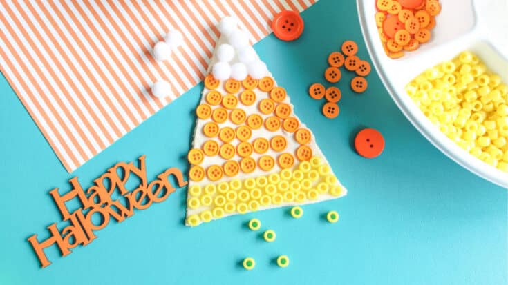 Candy corn playdough activity with a triangle of white, orange, and yellow playdough decorated with matching buttons and beads, next to a tray of craft supplies and a 'Happy Halloween' sign.