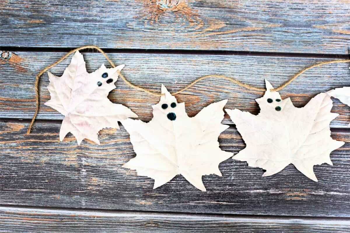 DIY ghost leaf garland made from painted white autumn leaves with black eyes and mouths, strung on twine against a rustic wooden background,