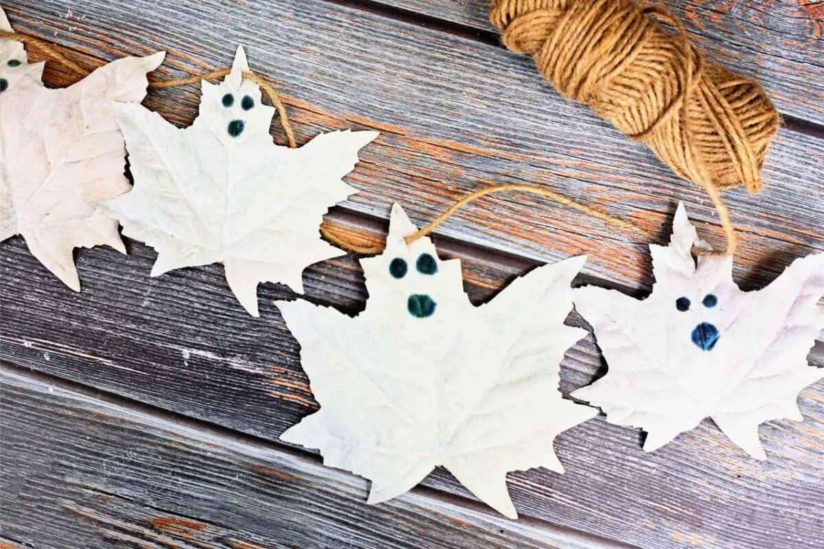 DIY ghost leaf garland made from painted white autumn leaves with black eyes and mouths, strung on twine against a rustic wooden background, Ball of twine in top right