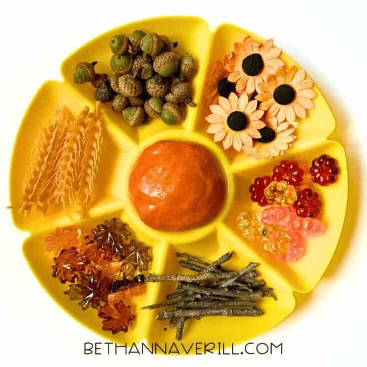 Yellow tray filled with autumn-themed playdough activity items including a ball of orange playdough in the center, surrounded by acorns, artificial sunflowers, red and yellow beads, twigs, plastic maple leaves, wheat stalks, and other loose parts.