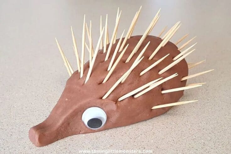 Brown playdough hedgehog craft with a googly eye and toothpicks for spikes on a light surface.