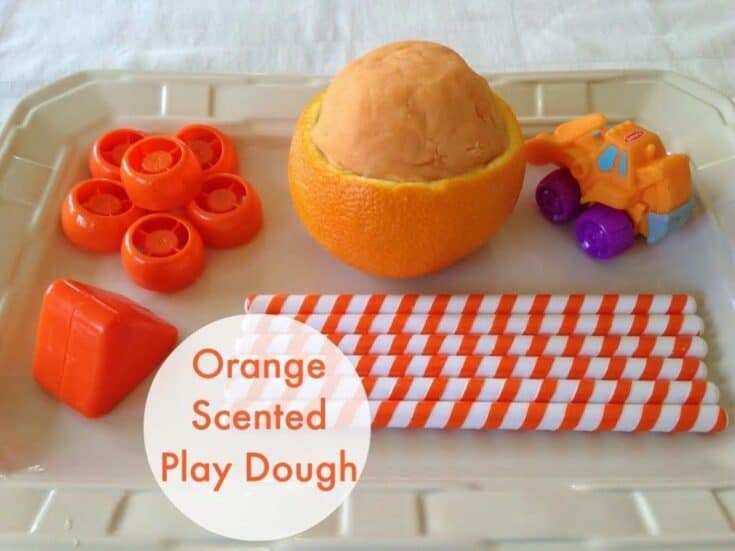 Tray with orange scented playdough inside a hollowed orange peel, toy truck, orange building blocks, orange wheels, and striped orange and white straws.