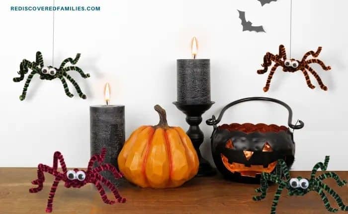 Halloween decorations with colorful pipe cleaner spiders with googly eyes placed around black candles, a pumpkin, and a jack-o’-lantern bucket