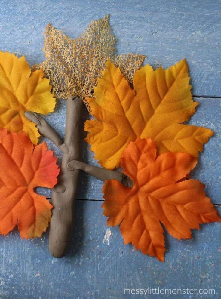 Children’s craft of an autumn tree made with brown playdough branches and orange and yellow fabric leaves on a blue wooden background.