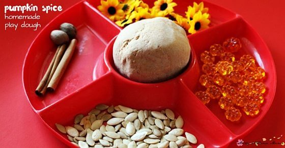 Red tray with sections containing a ball of homemade pumpkin spice playdough, pumpkin seeds, orange beads, yellow artificial flowers, cinnamon sticks, and nutmegs