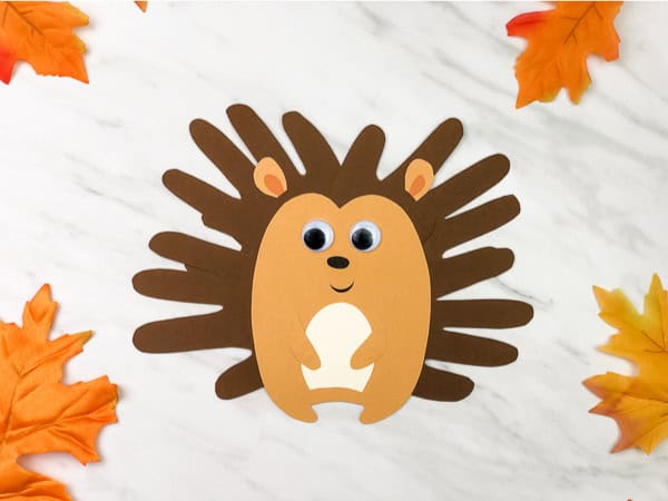 Handprint hedgehog craft for kids made from brown paper with googly eyes and autumn leaf decorations.