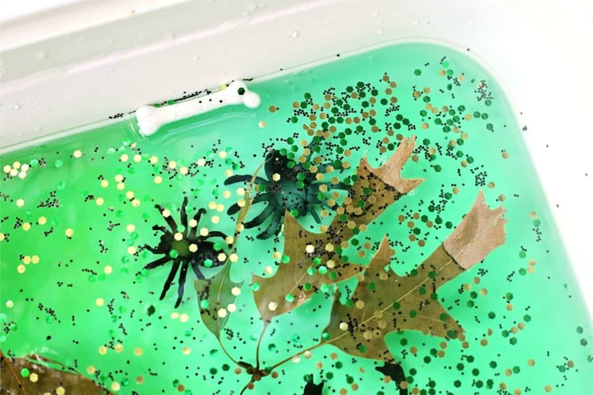 Close-up of a spooky swamp sensory bin with green water, gold glitter, toy spiders, plastic bones, and floating brown leaves