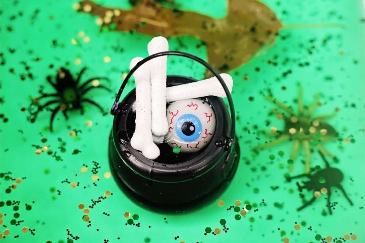 Black cauldron filled with toy bones and a plastic eyeball inside a spooky swamp sensory bin with green water, glitter, spiders, and leaves.