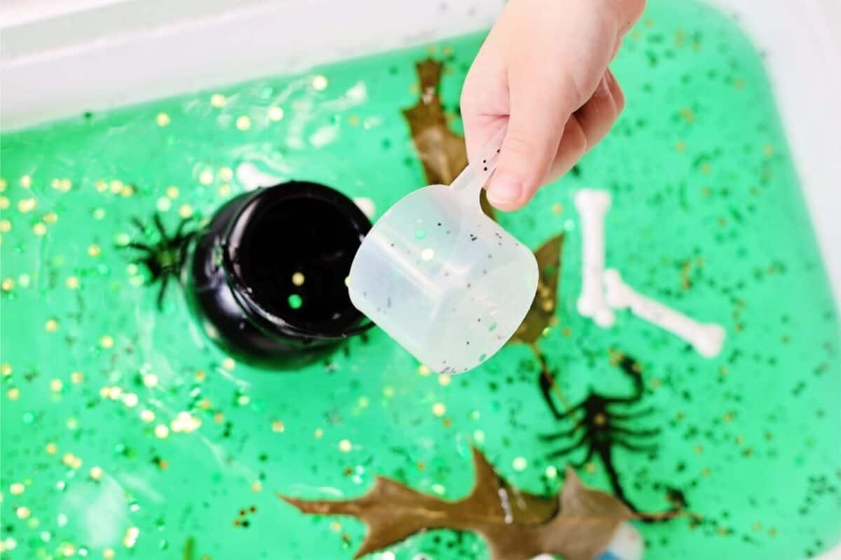 Child’s hand holding a plastic scoop over a spooky swamp sensory bin filled with green water, glitter, toy spiders, plastic bones, a black cauldron, and leaves.