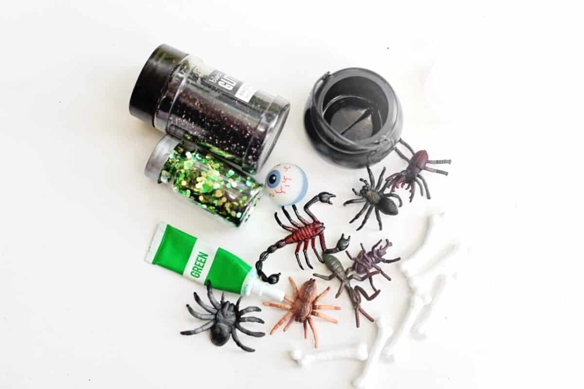 Supplies for a spooky swamp sensory bin including green paint, black and green glitter, toy spiders, scorpions, bones, a plastic eyeball, and a black cauldron.