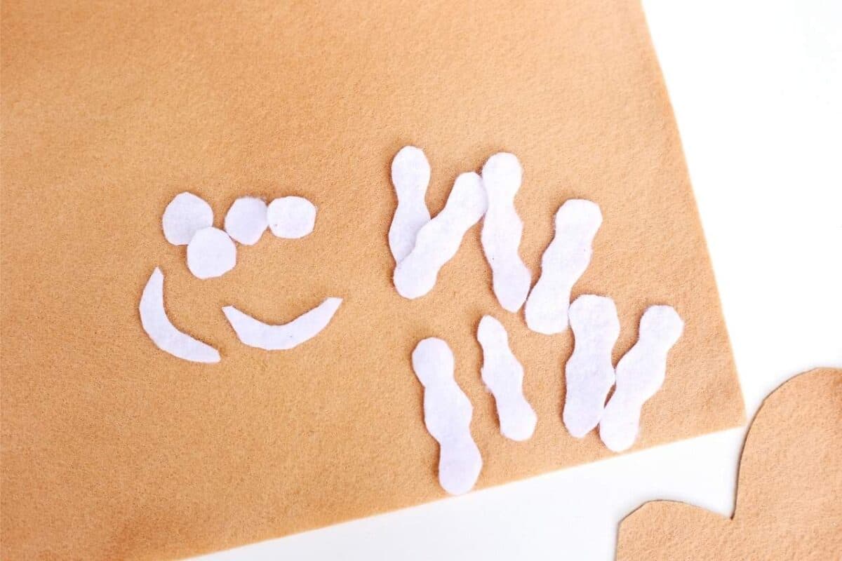 White felt pieces cut out for gingerbread decorations including eyes, smiles, and icing strips placed on tan felt.