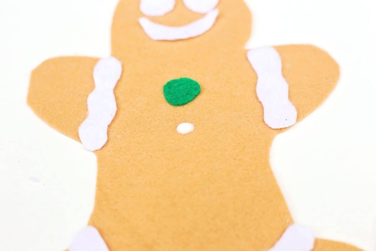 Close up of a felt gingerbread person with white icing strips and green button being glued in place.