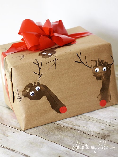 A wrapped gift in brown paper decorated with painted brown reindeer footprints featuring googly eyes, antlers, and red noses. The gift is topped with a large red bow.