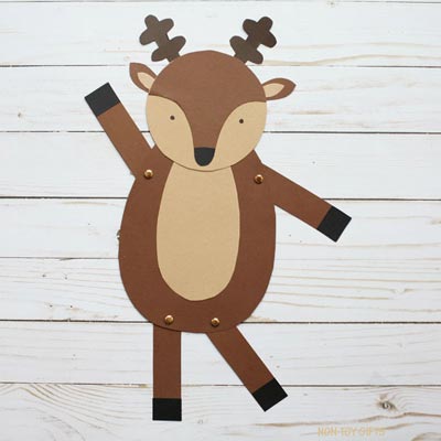 A flat paper reindeer craft made from brown and beige cardstock, laid on a white wooden surface. The reindeer has movable arms and legs attached with brass fasteners, small black hooves, a beige belly, and simple cut-out antlers and ears.