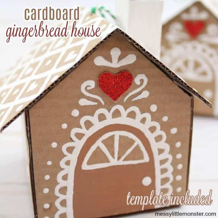 Cardboard gingerbread house craft decorated with white painted icing-style patterns and a red glitter heart above the door, made from recycled cardboard with a printed template.