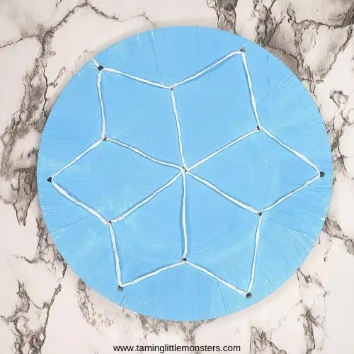 Circular cardboard lacing craft painted light blue with a six-point star flower shape stitched in white yarn through punched holes. The project is placed on a white and gray marble background.