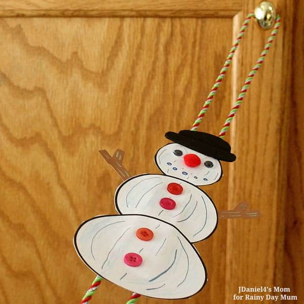 Handmade snowman craft with three white paper circles outlined in black, decorated with red and pink buttons, a black hat, and a red pom-pom nose. The snowman is attached to colorful twisted yarn, allowing it to slide or hang from a doorknob.