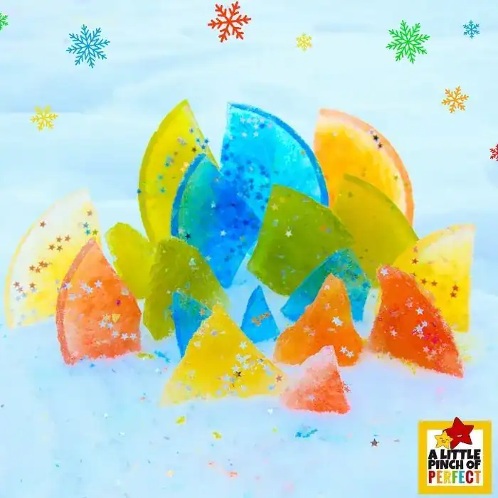 Brightly colored frozen ice shapes in yellow, blue, green, and orange arranged upright in snow to resemble a sculpture or cityscape. Each piece is sprinkled with glitter and star-shaped confetti, adding a festive, artistic touch. Snowflakes and the logo “A Little Pinch of Perfect” decorate the background.