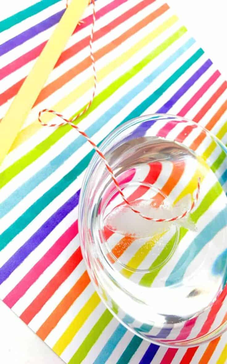Clear glass of water with a melting ice cube and a red-and-white striped string partially frozen to the ice, sitting on a brightly colored rainbow-striped paper. This setup is part of a classic ice and salt STEM experiment to lift ice with a string.